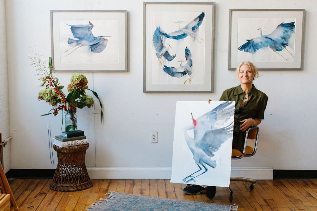 Holly Wach (artist)  sitting in a chair holding her original bird watercolor art